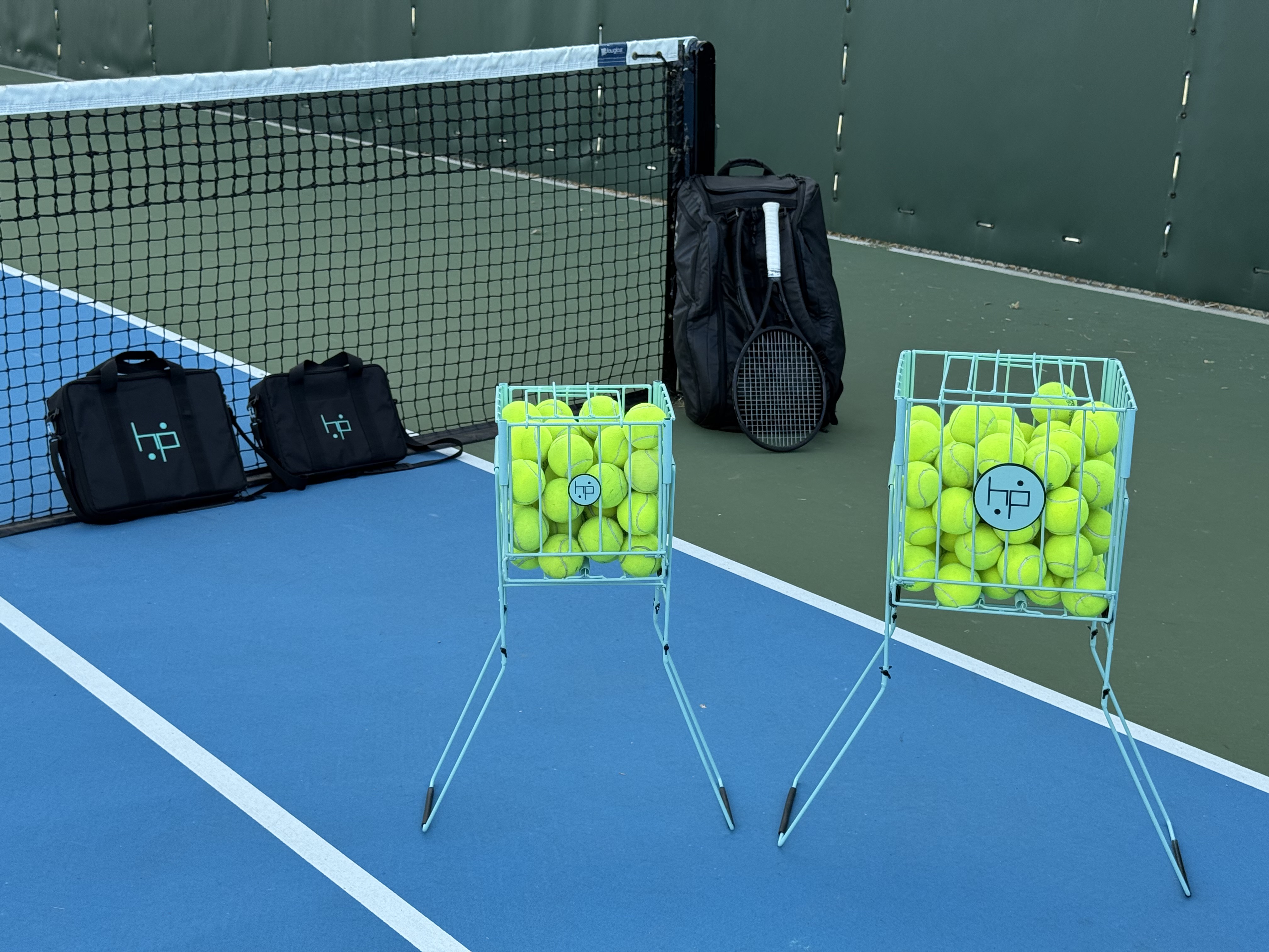 Two mint BallHP foldable hoppers filled with tennis balls beside the net on a blue hard court